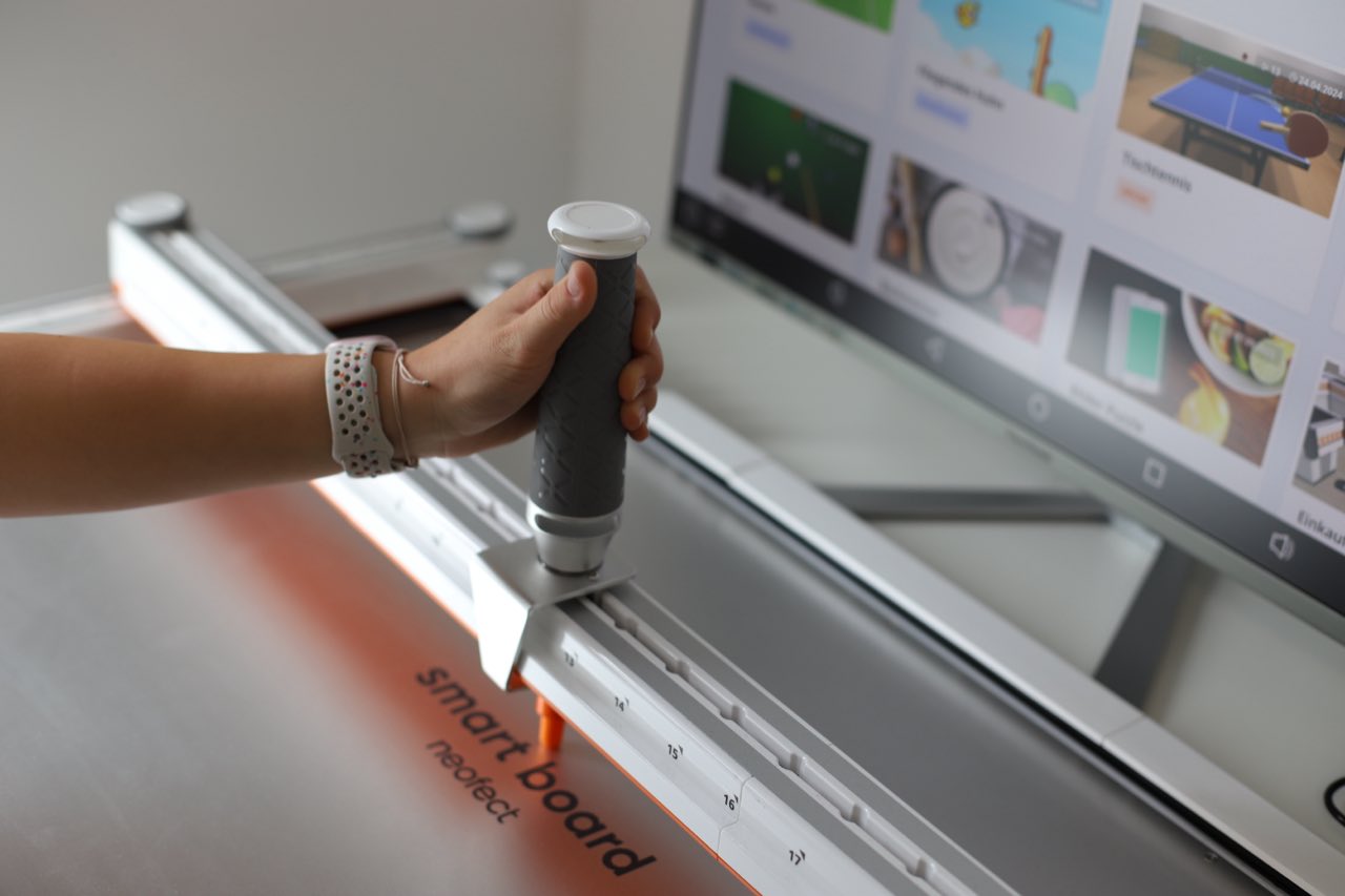 Eine Hand führt den Griff des Neofect Smart Board, auf dem Bildschirm dahinter laufen Übungsspiele